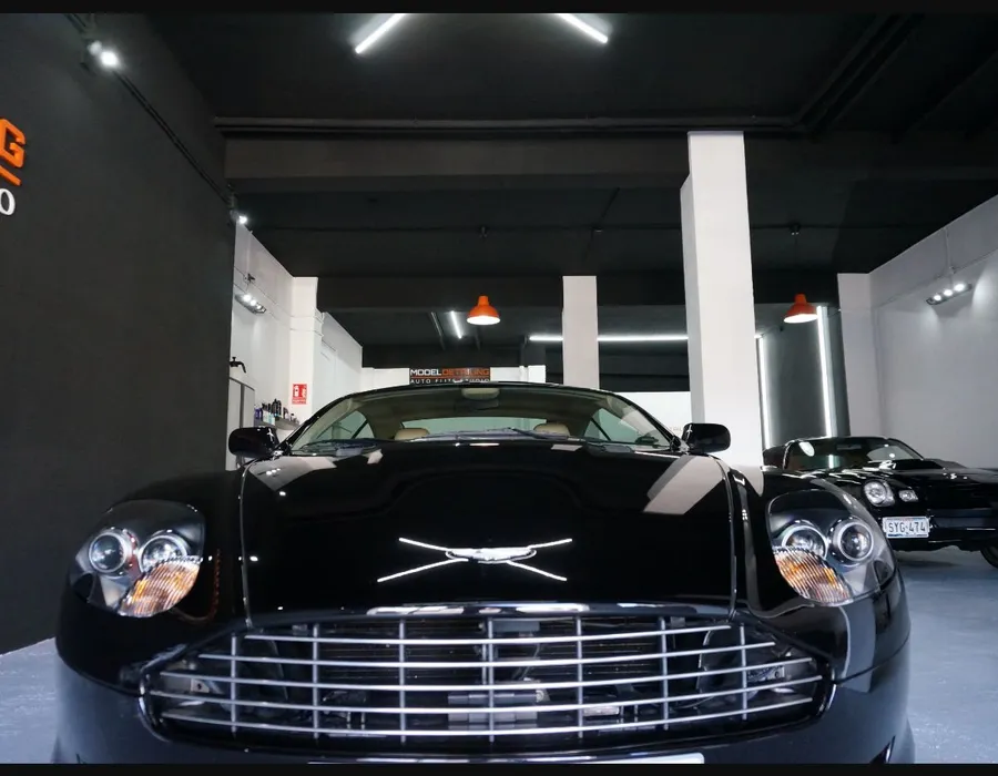 Aston Martin negro fotografiado de frente en el estudio, con otros vehículos clásicos al fondo.