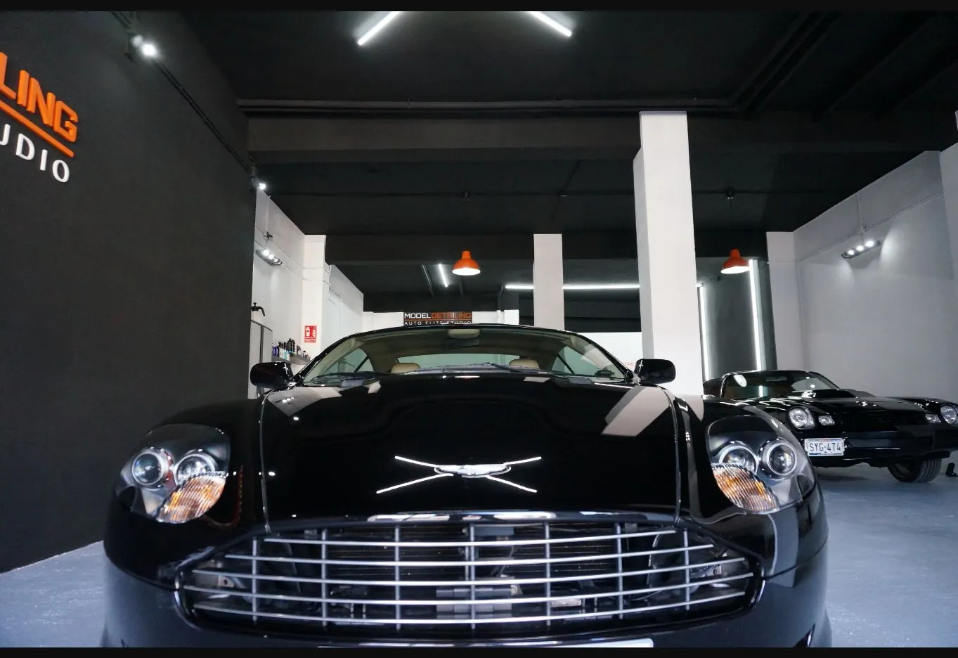 Aston Martin negro fotografiado de frente en el estudio de Model Detailing, con otros vehículos clásicos al fondo.